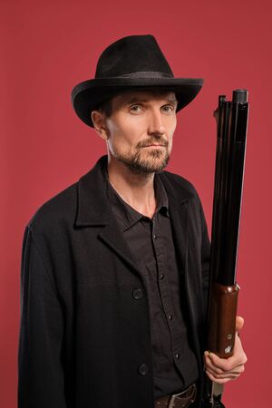 Middle-aged man with beard, mustache, in black jacket and hat, holding a gun while posing against a...