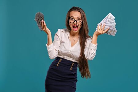 Brunette girl in glasses, wearing in a black short skirt and white blouse is posing holding a fan of hundred dollar bills against a blue background.の写真素材