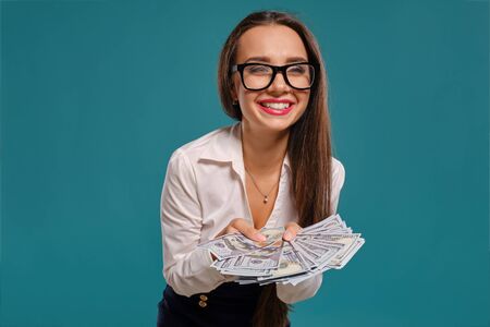 Brunette girl in glasses, wearing in a black short skirt and white blouse is posing holding a fan of hundred dollar bills against a blue background.の写真素材