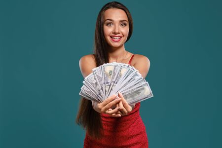 Brunette girl with a long hair, wearing a sexy red dress is posing holding a fan of hundred dollar bills against a blue background.の写真素材