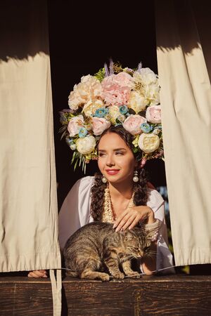 Brunette girl in a white ukrainian authentic national costume and a wreath of flowers is is looking out of the window. Close-up.の写真素材