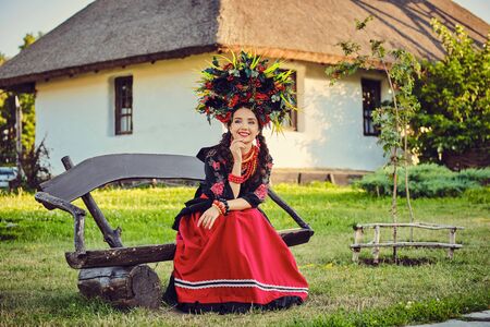 Brunette girl in a white ukrainian authentic national costume and a wreath of flowers is posing sitting on a wooden bench against a white hut.の写真素材