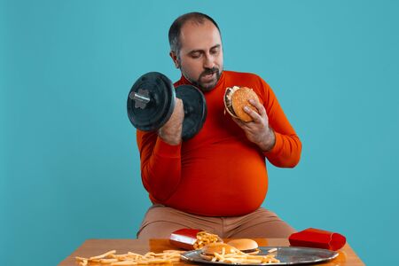 Close-up portrait of a middle-aged man with beard, dressed in a red turtleneck, posing with burgers and french fries. Blue background. Fast food.の写真素材