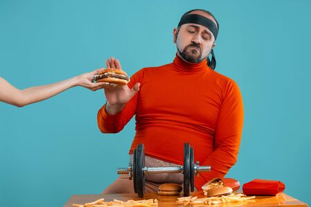 Middle-aged man with beard, dressed in a red turtleneck, headband, posing with burgers and french fries. Blue background. Close-up. Fast food.の写真素材