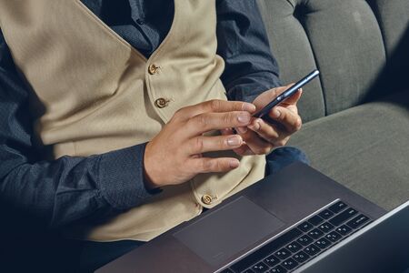 Hands of a man, dressed in black shirt and beige vest is sitting on dark sofa, using his laptop and...