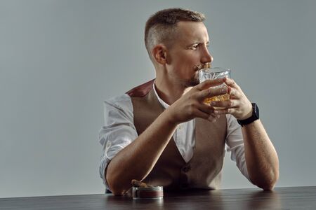 Man with stylish mustache, dressed in classic brown vest, white shirt is sitting at table, enjoying...