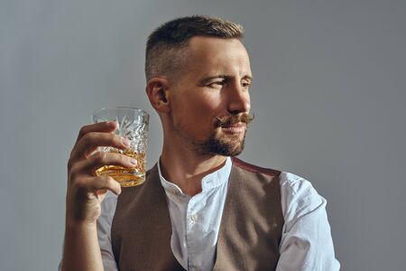 Man with stylish mustache, dressed in classic brown vest, white shirt is sitting at the table, enjoying whiskey. Grey background, close-up shot.の写真素材