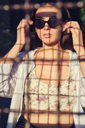 Portrait of girl in sunglasses posing in city behind a trellised fence. Dressed in top with floral...