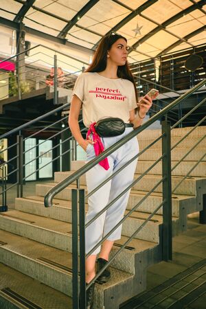 Portrait of a girl posing near a shopping mall, holding smartphone. Dressed in white t-shirt, blue...