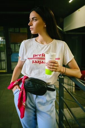 Portrait of girl with dark sunglasses on her head posing in shopping mall, holding drink. Dressed in...
