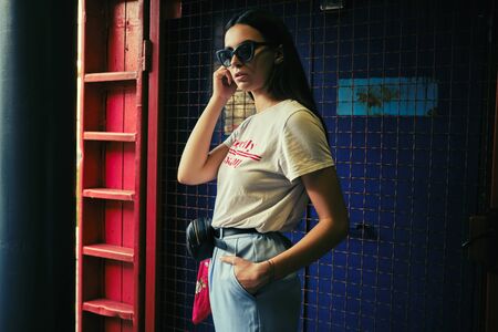 Portrait of a girl in dark sunglasses posing at a blue gate near the mall. Dressed in white t-shirt,...