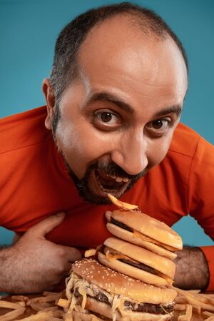 Close-up portrait of a middle-aged man with beard, dressed in a red turtleneck, posing with burgers and french fries. Blue background. Fast food.の写真素材