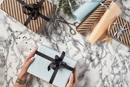 Hands of a woman are holding white paper gift box tied with black ribbon and bow against a marble background. Wrapping paper, decorative little stars, green twig and scissors are laying nearby. Concept of holidays, fests, celebrations, congratulations, presents, decorations, greetings. Close-up shot. Copy space. Top view.の写真素材