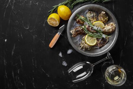 Fresh yummy closed oysters, ice, lemon and a green twig of lavender on a round metal plate and two glasses of white wine are on a black stone textured background. Oyster knife is laying nearby. Supper at a restaurant. Healthy sea food. Top view with copy space. Close-up shot.の写真素材