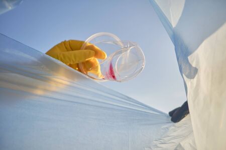 Hand of a conscious human in a yellow rubber glove puts a rumpled plastic cup in a blue trash bag. Blue sky. People and ecology. Riverside pollution. Preservation of nature. Volunteering concept. Close-up shot.の写真素材