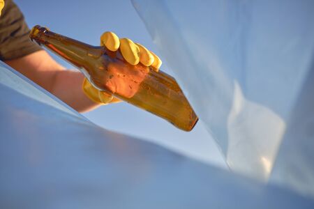 Hand of a woman in a yellow rubber glove puts a used brown glass bottle in a blue trash bag. Blue sky. People and ecology. Riverside pollution. Preservation of nature. Volunteering concept. Close-up shot.の写真素材