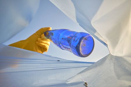 Hand of a conscious person in a yellow rubber glove puts a used blue glass bottle in a trash bag. Blue sky. People and ecology. Riverside pollution. Preservation of nature. Volunteering concept. Close-up shot.の写真素材