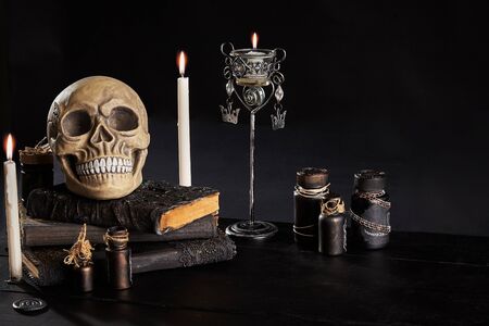 Realistic model of a human cranium with teeth is standing on an old books, jars, candlestick and burning candles are nearby on a wooden dark table. Black background. Medical science or Halloween horror concept. Magic, wizardry, miracles. Close-up shot.の写真素材