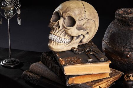 Realistic model of a human skull with teeth is standing on an old books against a dark pot and candle in candlestick. Black background. Medical science or Halloween horror concept. Magic, wizardry, miracles. Close-up shot.の写真素材