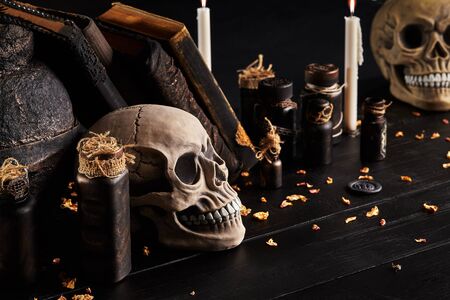 Realistic model of two human skulls with white teeth, burning candles, books with spells, old pot and jars of potion are on a wooden dark table. The petals of dried roses are scattered nearby. Black background. Medical science or Halloween horror concept. Magic, wizardry, miracles. Close-up shot. Side view.の写真素材