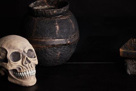 Realistic model of a human skull with teeth, old pot and books are on a wooden dark table. Black background. Medical science or Halloween horror concept. Magic, wizardry, miracles. Close-up shot.の写真素材