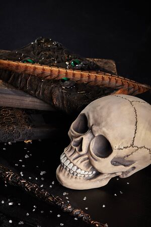 Realistic model of a human cranium with teeth, books, feather, magic wand are on a wooden dark table. Small broken glass is scattered around. Black background. Medical science or Halloween horror concept. Magic, wizardry, miracles. Close-up top view.の写真素材