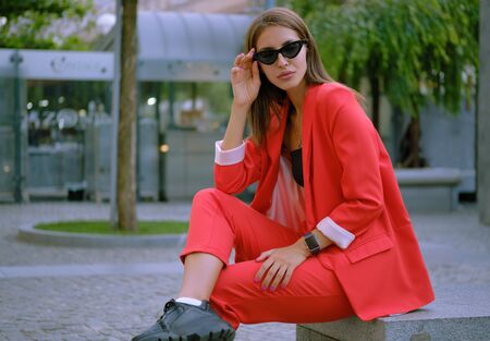 Blonde girl in a red lady-type pantsuit and black top, watch, ring, pendant and sunglasses is posing sitting on a stone bench. Close-up shot.の写真素材