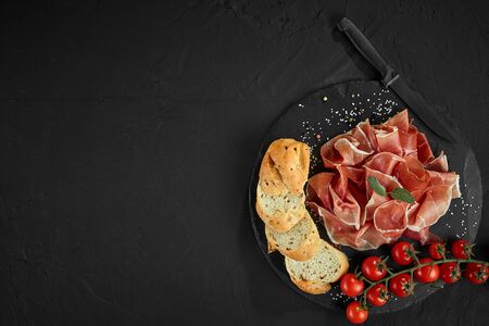Sliced jamon, cherry tomatoes, baguette, herbs, spices and knife on black stone slate board against...