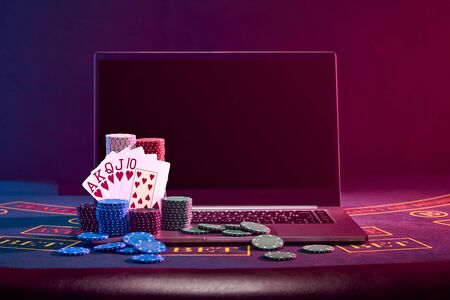 Chips piles and playing cards on laptop standing on blue cover of a table. Black background....