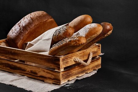Assortment of fresh baked bread in a paper bag and wooden box standing on a burlap against black...