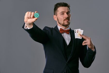Man in black classic suit and red bow-tie showing two playing cards and chip, posing on gray studio...
