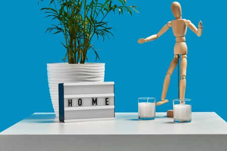 White coffee table with green flower in pot, two candles, wooden figurine of human and decor element with inscription home. Blue background. Close upの写真素材