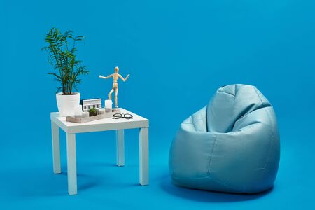 Turquoise bean bag, white coffee table with green flower in pot, candles, succulent, wooden figurine of human, glasses. Blue background. Copy spaceの写真素材