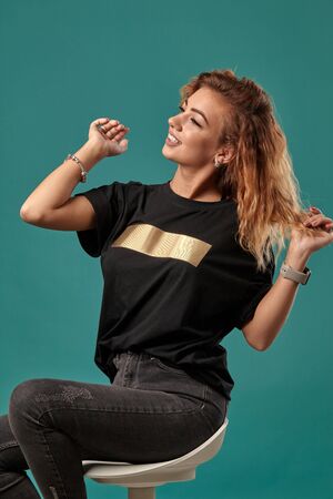 Ginger girl with a curly hair, wearing black t-shirt with a golden stripe is posing sitting on a...