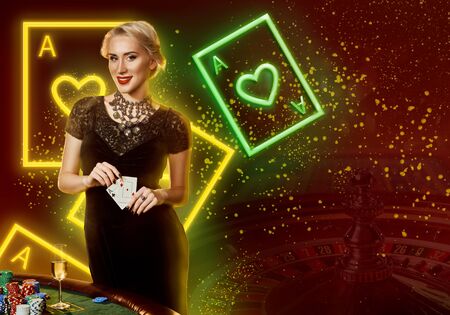 Girl in black dress showing aces, smiling, posing at table with chips, champagne. Colorful background with neon playing cards, roulette. Poker, casinoの写真素材