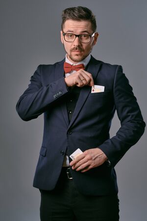 Man in black classic suit, red bow-tie, glases is showing two playing cards, posing on gray studio background. Gambling, poker, casino. Close-up.の写真素材