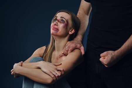 Strong man put his hand on female shoulder, showing fist. Victim with bruises on face sitting...