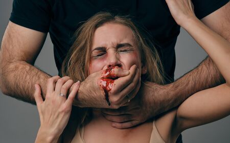 Strong man strangling scared female with bruises on her face. Blood from her mouth flows down his arm. Gray background. Domestic violence. Close-up.の写真素材