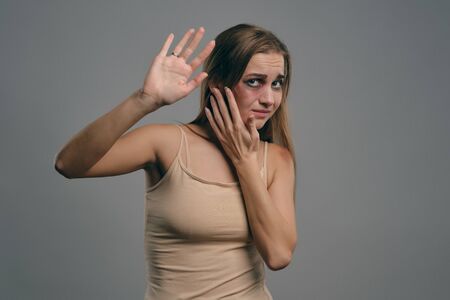Blonde girl with bruises on her face, trying to protect herself, posing on gray studio background....