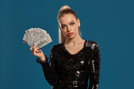 Blonde lady with ponytail, in black sequin dress. Showing fan of hundred dollar bills, posing on...