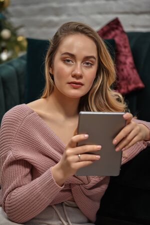 Female blogger in pink sweater. Sitting leaning on gray sofa and holding her tablet. Interior with...