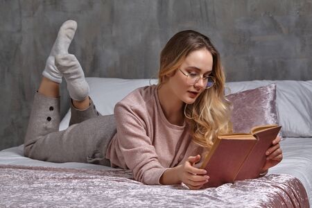 Blonde female in glasses, casual clothing. She is laying on bed and reading book in bedroom....
