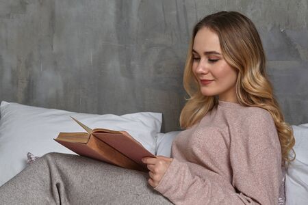 Female in casual clothing. Smiling, laying on bed, reading book in bedroom. Student, blogger,...