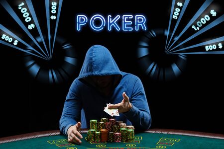 Man in blue hoodie with hood is showing two aces, posing sitting at a green playing table with...