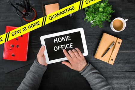 Quarantine, stay home. Coronavirus, yellow warning tape. Top view. Hands of man typing on tablet, home office inscription on screen. Close upの写真素材