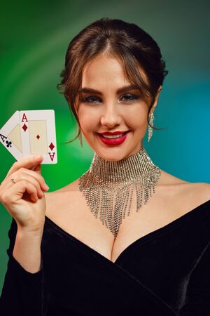 Brunette model in black dress, necklace and earrings. Smiling, showing two playing cards, posing on colorful background. Poker, casino. Close-upの写真素材