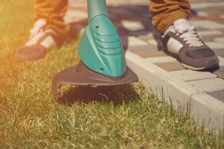 Male in sneakers and pants is cutting green grass with handheld modern lawn mower in his garden....