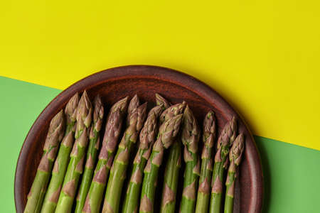 Asparagus stalks in brown clay plate, over yellow and green background. Concept of food and seasonal vegetables harvest. Close up, copy spaceの写真素材