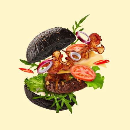 Fresh black burger with flying ingredients on beige background. Ham, beef cutlet, cheese, sauces, vegetables and greens. Close up, copy spaceの写真素材