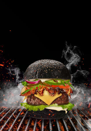 Black burger roasted on barbecue grill with flaming fire, sparks and smoke against black background. Beef cutlet, ham, cheese, vegetables and greensの写真素材